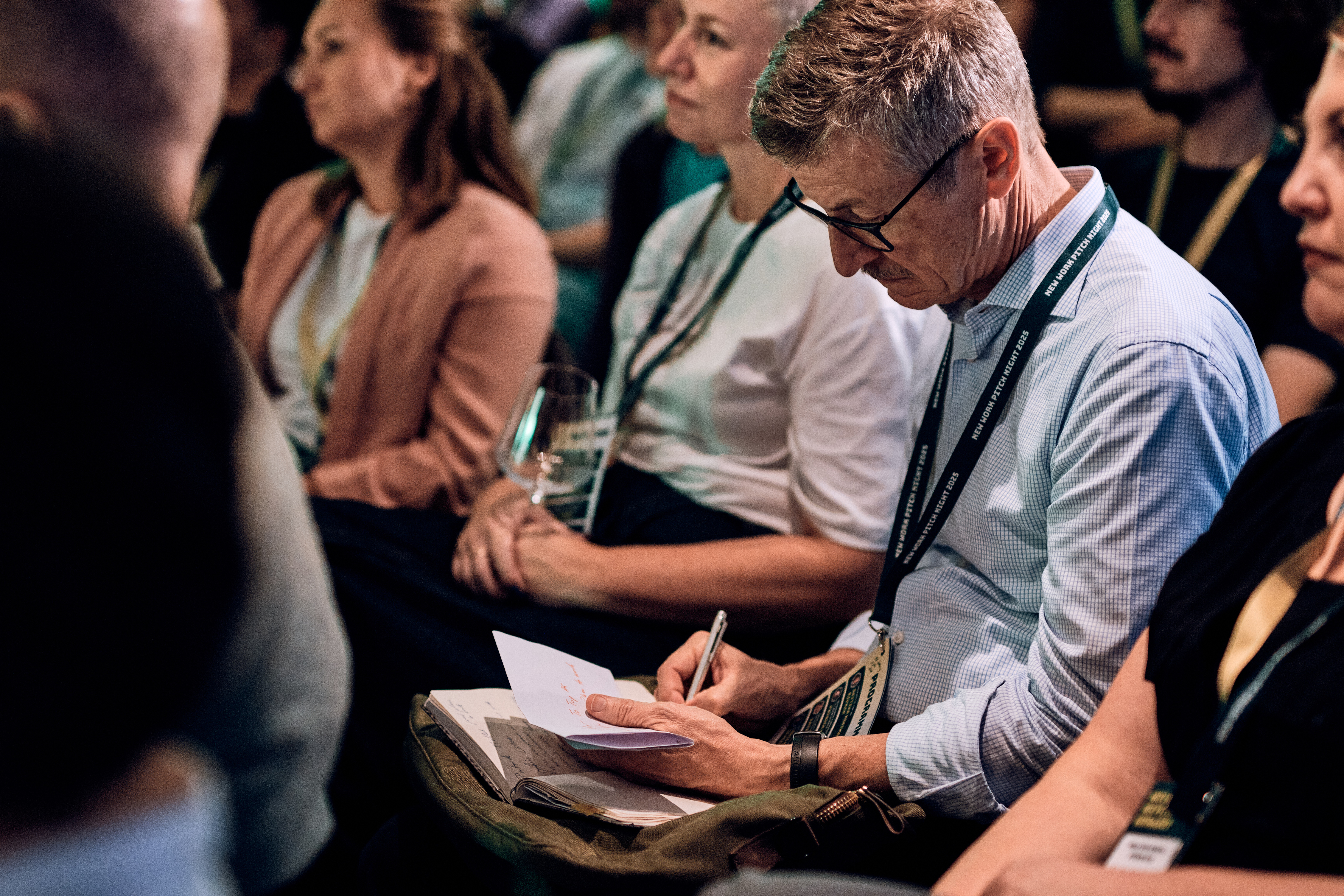 Ein Mann mit Brille sitzt inmitten anderer Personen und macht sich Notizen in einem Notizbuch, das wie ein Seminar oder eine Konferenz aussieht.